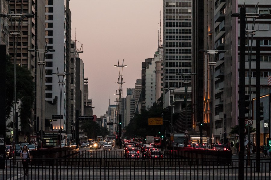 Eixo da Avenida Paulista, em São Paulo, ao entardecer, com a fileira de postes e o céu quente.